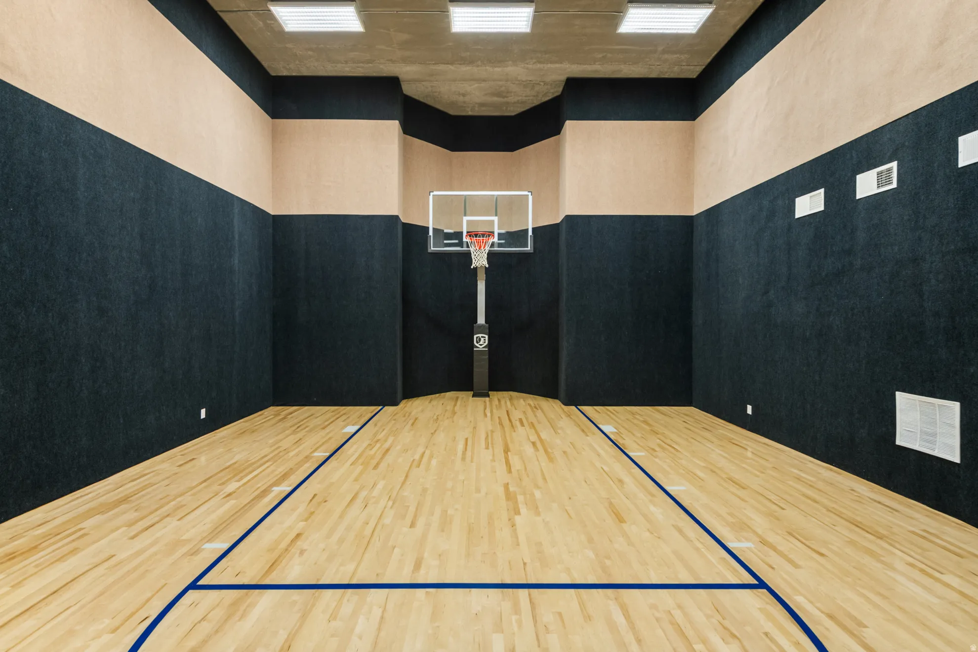 Indoor court featuring wood-finish flooring with blue demarcation lines, a basketball hoop with a glass backboard, sound-dampening wall panels, and recessed ceiling lighting