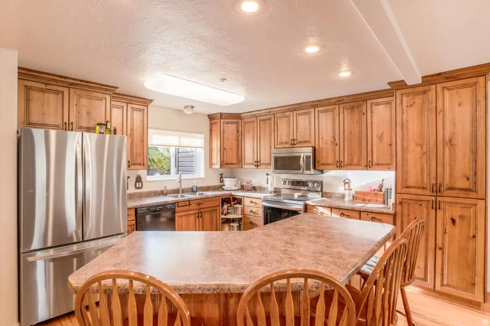 Kitchen featuring wood cabinetry, stainless steel appliances, a corner sink with window, recessed lighting, and a breakfast bar with stone-finish countertop