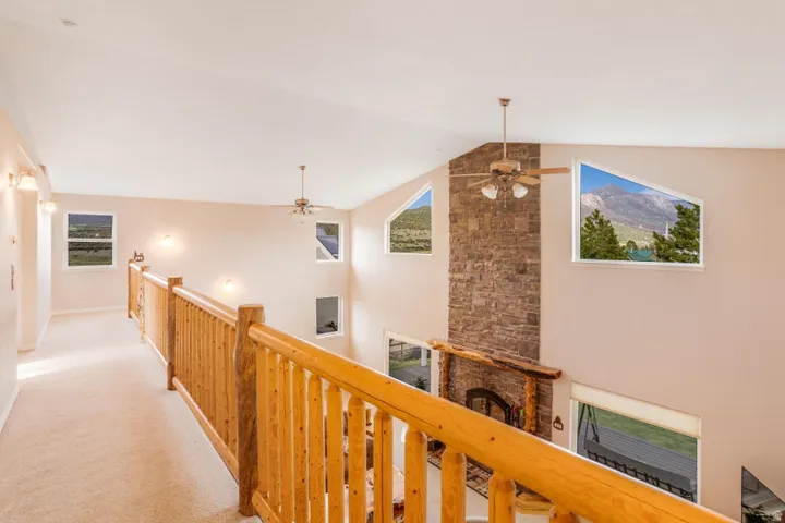 Upper-level hallway overlooking a great room with a stone fireplace, exposed wood mantel, and ceiling fan