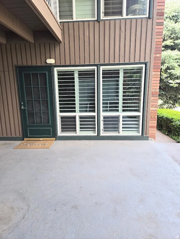 Patio entrance featuring a dark green paneled door with glass inserts, adjacent to two large windows fitted with white plantation shutters