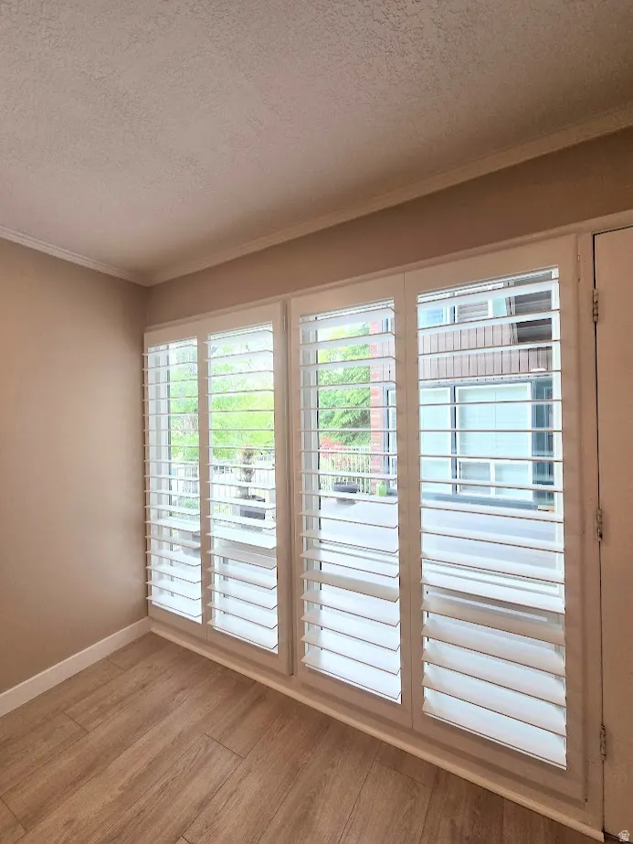 Triple window unit featuring white plantation shutters, wood-finish flooring, crown molding, and neutral wall tones