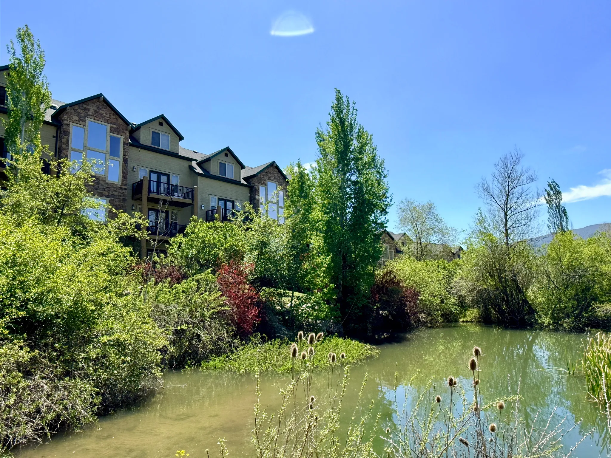 Pond-side residence featuring multi-level balconies, stone accents, dormer windows, and surrounding natural landscaping