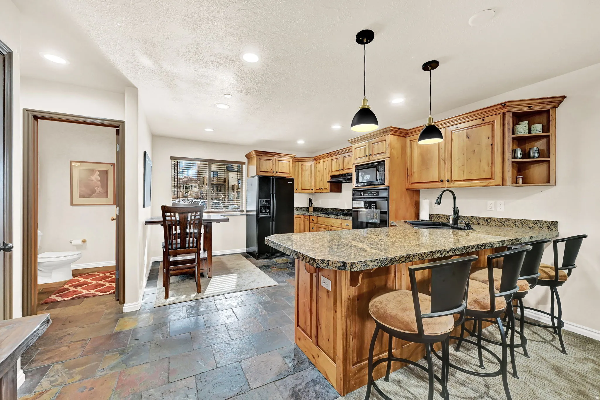 Kitchen with wood cabinetry, stone countertops, integrated black appliances, a counter-height breakfast bar, and slate-style flooring