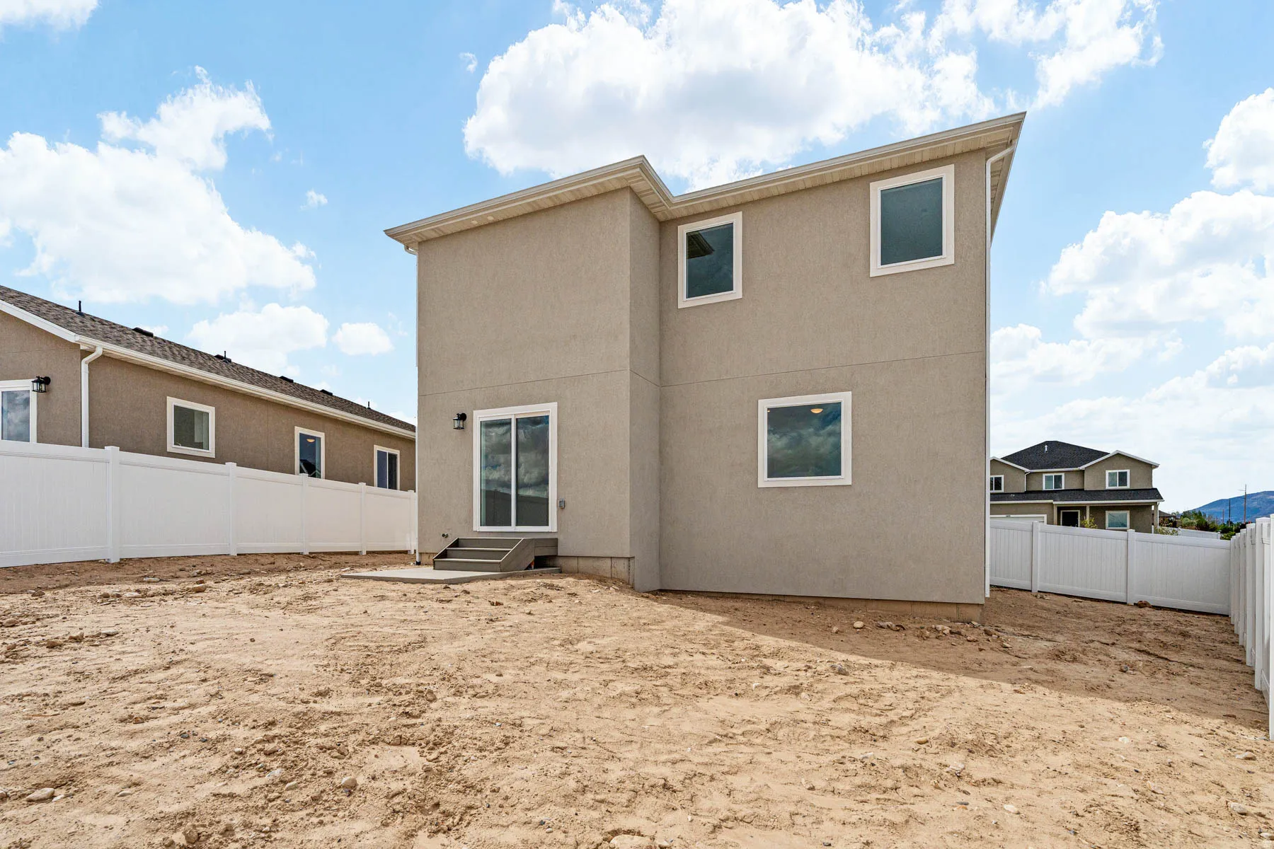 Rear exterior with a stucco finish, showcasing multiple windows, a sliding glass door, a concrete patio, and a white privacy fence