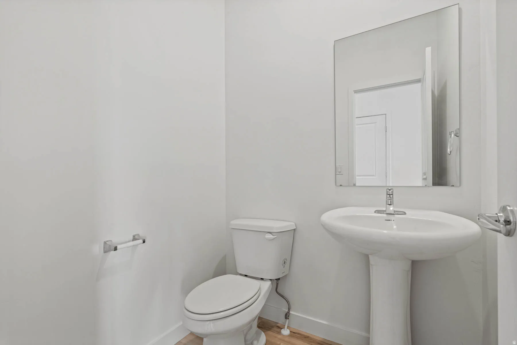 Bathroom featuring a white pedestal sink, coordinating toilet, a frameless wall mirror, and wood-finish flooring