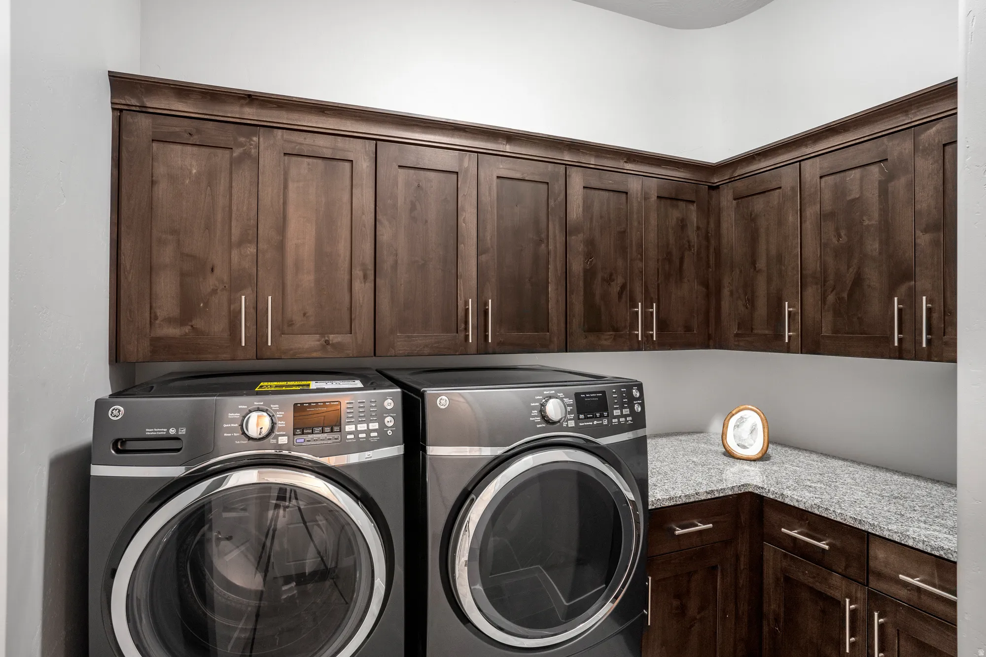 Laundry room featuring wood-finish cabinetry with silver hardware, light-toned granite-style countertops, and built-in upper and lower storage