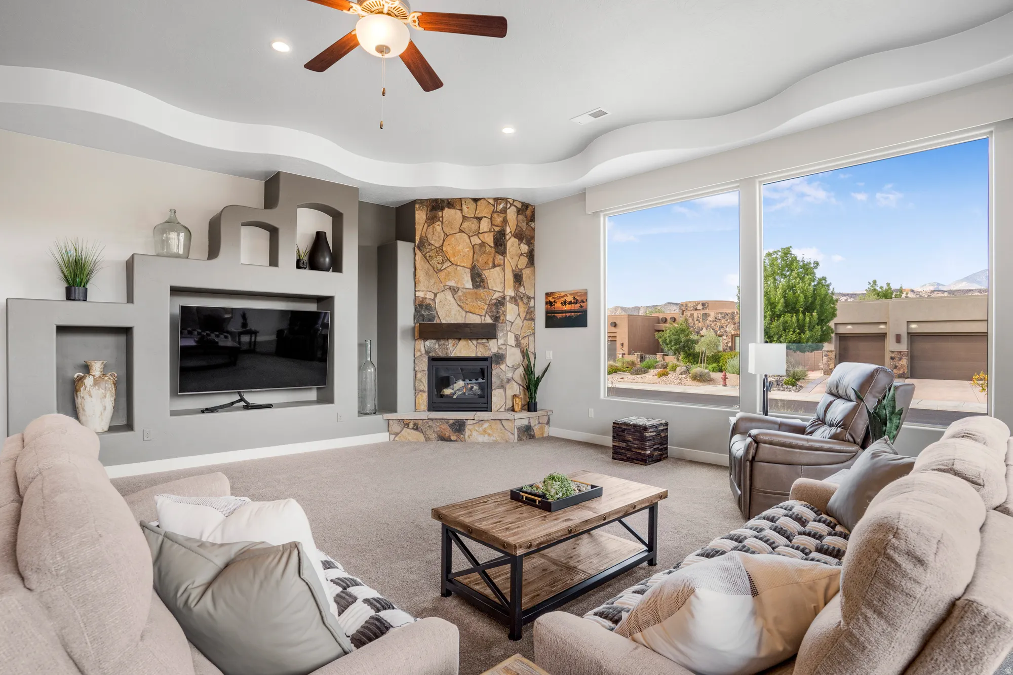 Living area featuring large windows, a stone fireplace, a built-in entertainment center with shelving, a tray ceiling, and carpeted flooring
