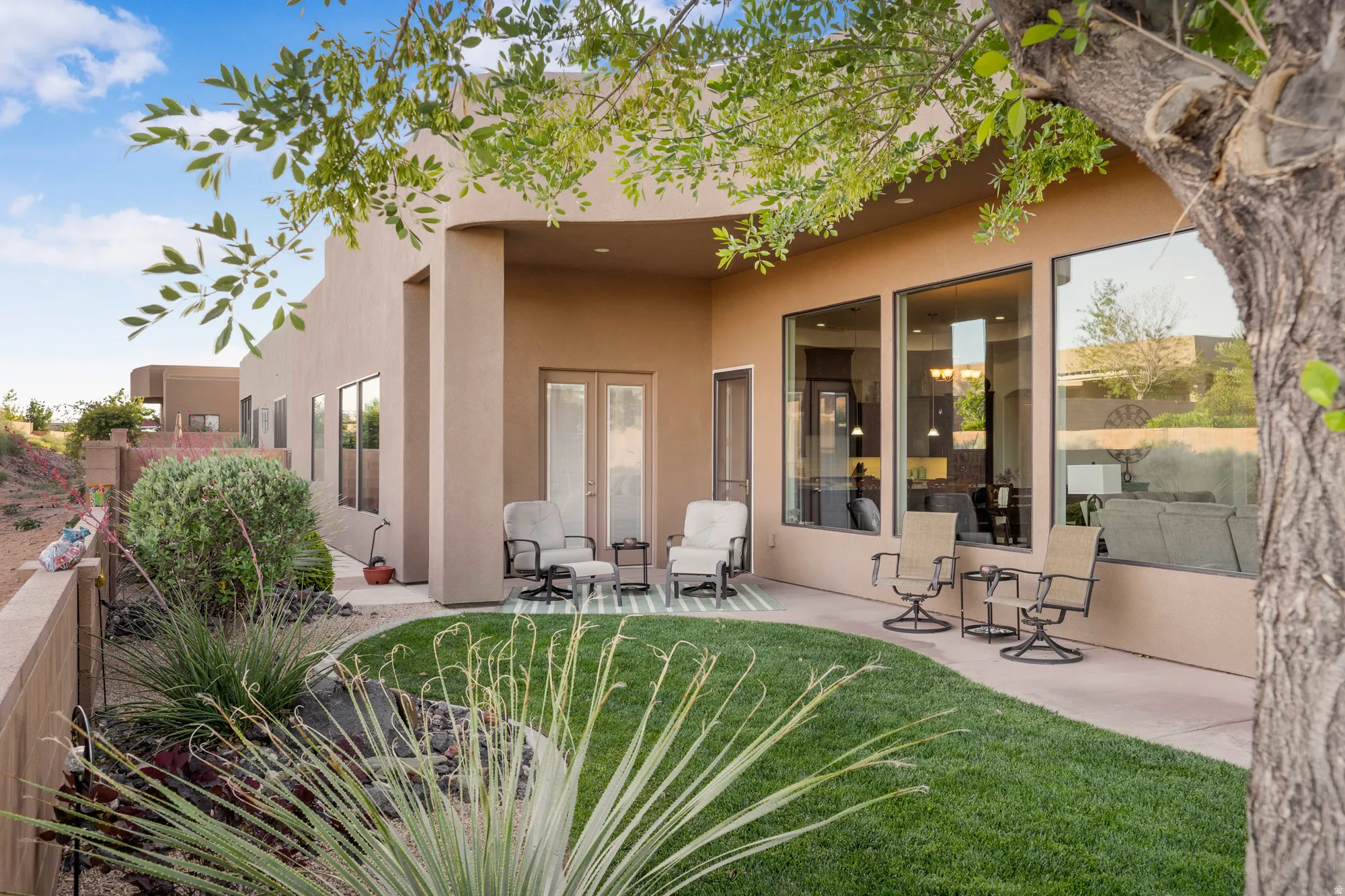 Expansive outdoor living space featuring a curved green lawn, mature desert landscaping, a covered patio, a privacy wall, and large windows with integrated glass doors