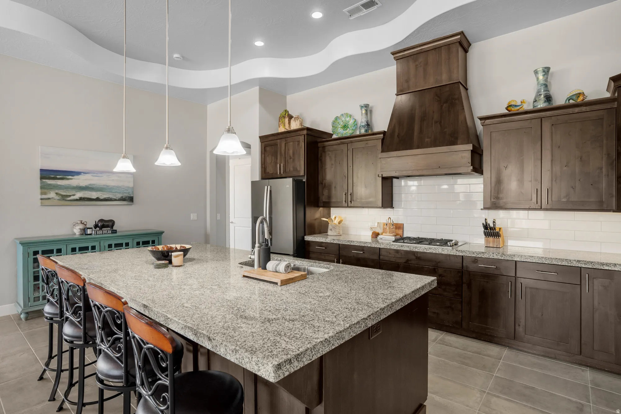 Kitchen featuring an expansive island with a granite countertop, dark wood-finish cabinetry, a built-in gas range, a coordinating wood-finish range hood, and a subway tile backsplash