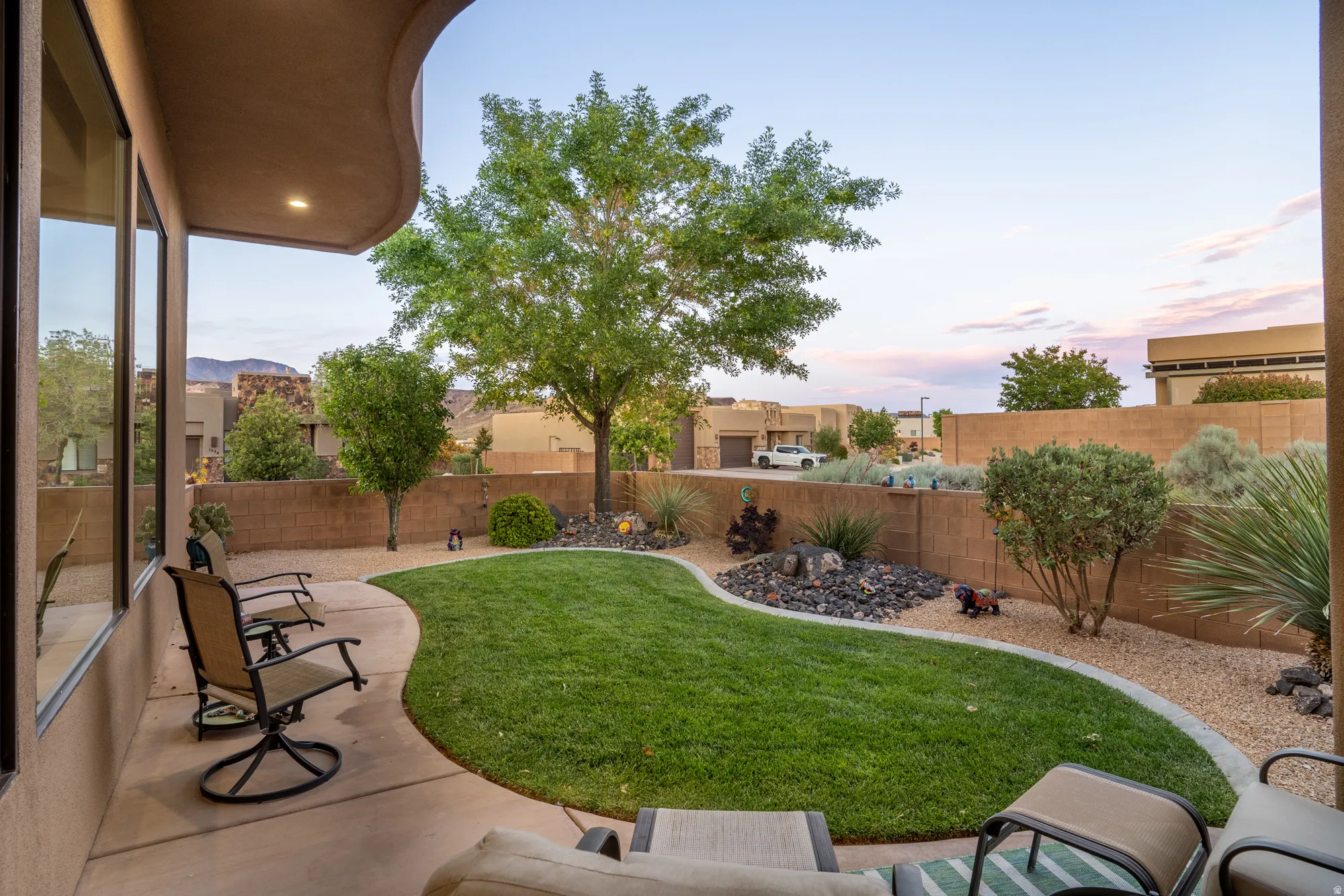 Covered patio overlooking a landscaped backyard featuring a curved lawn, gravel beds, mature trees, and a block fence