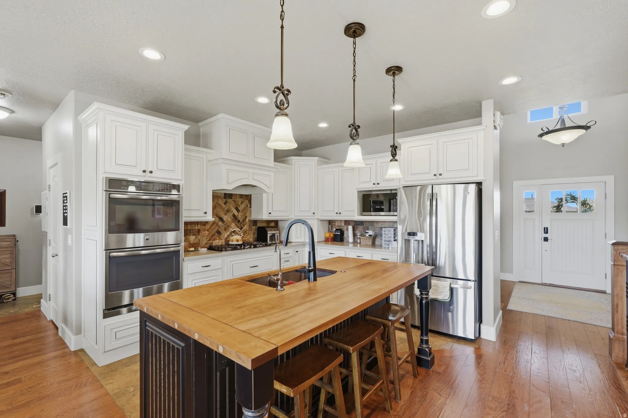 Kitchen featuring a central island with a wood countertop and integrated sink, white cabinetry with dark hardware, stainless steel double wall ovens, a gas range with a decorative hood, and hardwood flooring