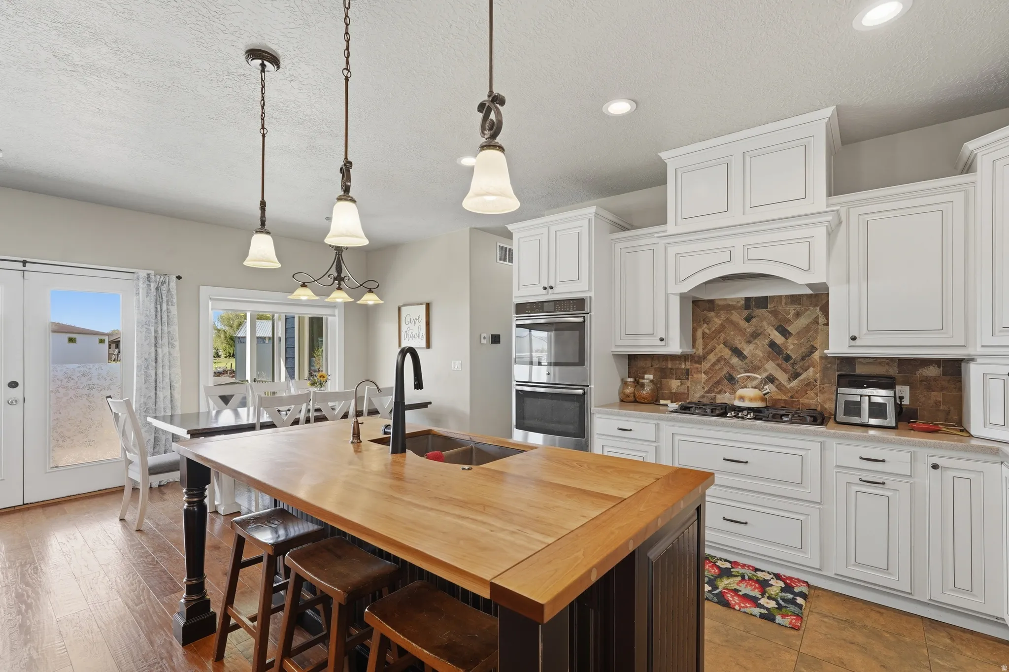Spacious kitchen featuring an island with butcher block countertop, white cabinetry with dark hardware, a herringbone pattern tile backsplash, double wall ovens, and wood-finish flooring
