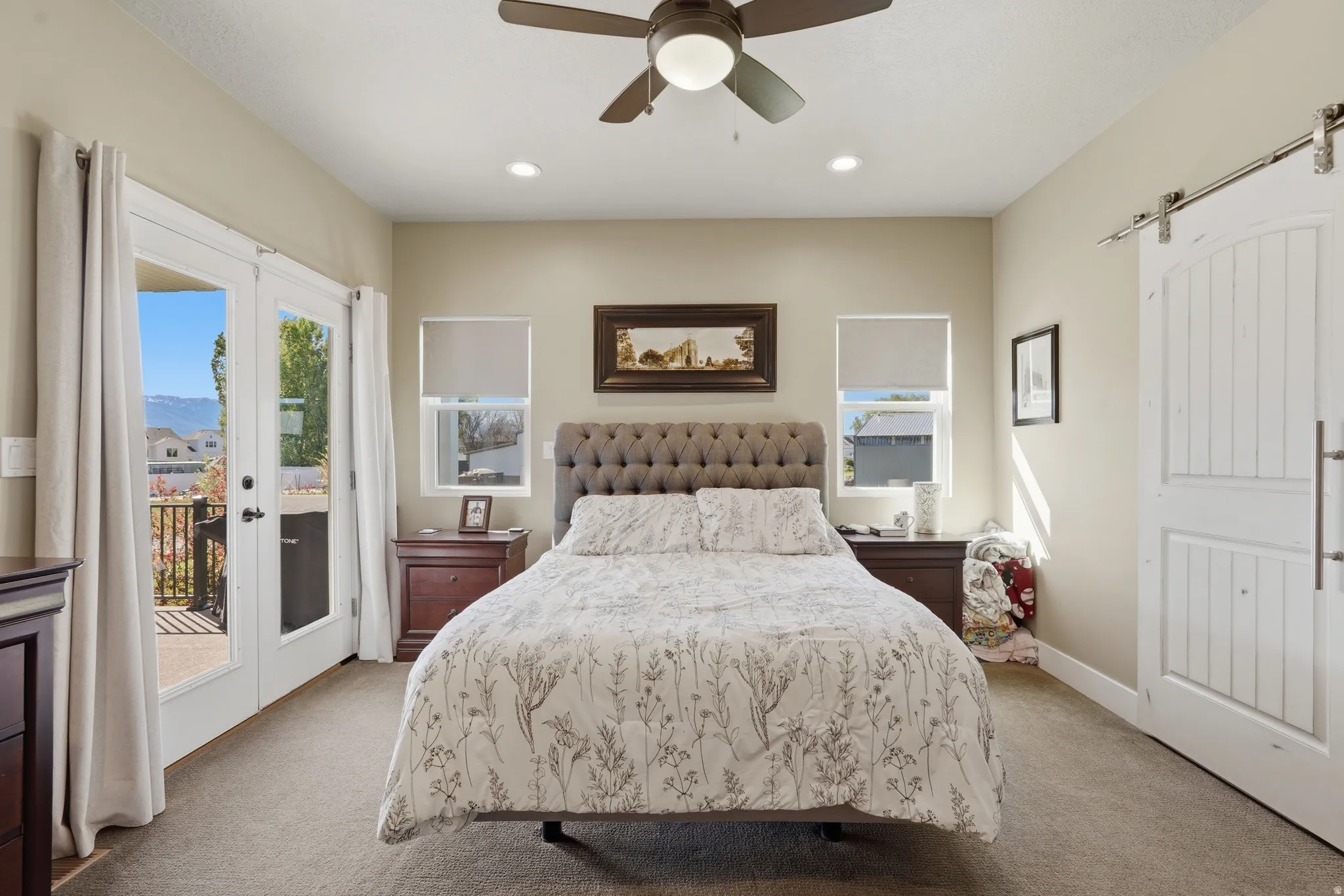 Plush carpeted bedroom featuring double French doors leading to an exterior balcony, two windows with roller shades, a ceiling fan with integrated lighting, and a white barn door
