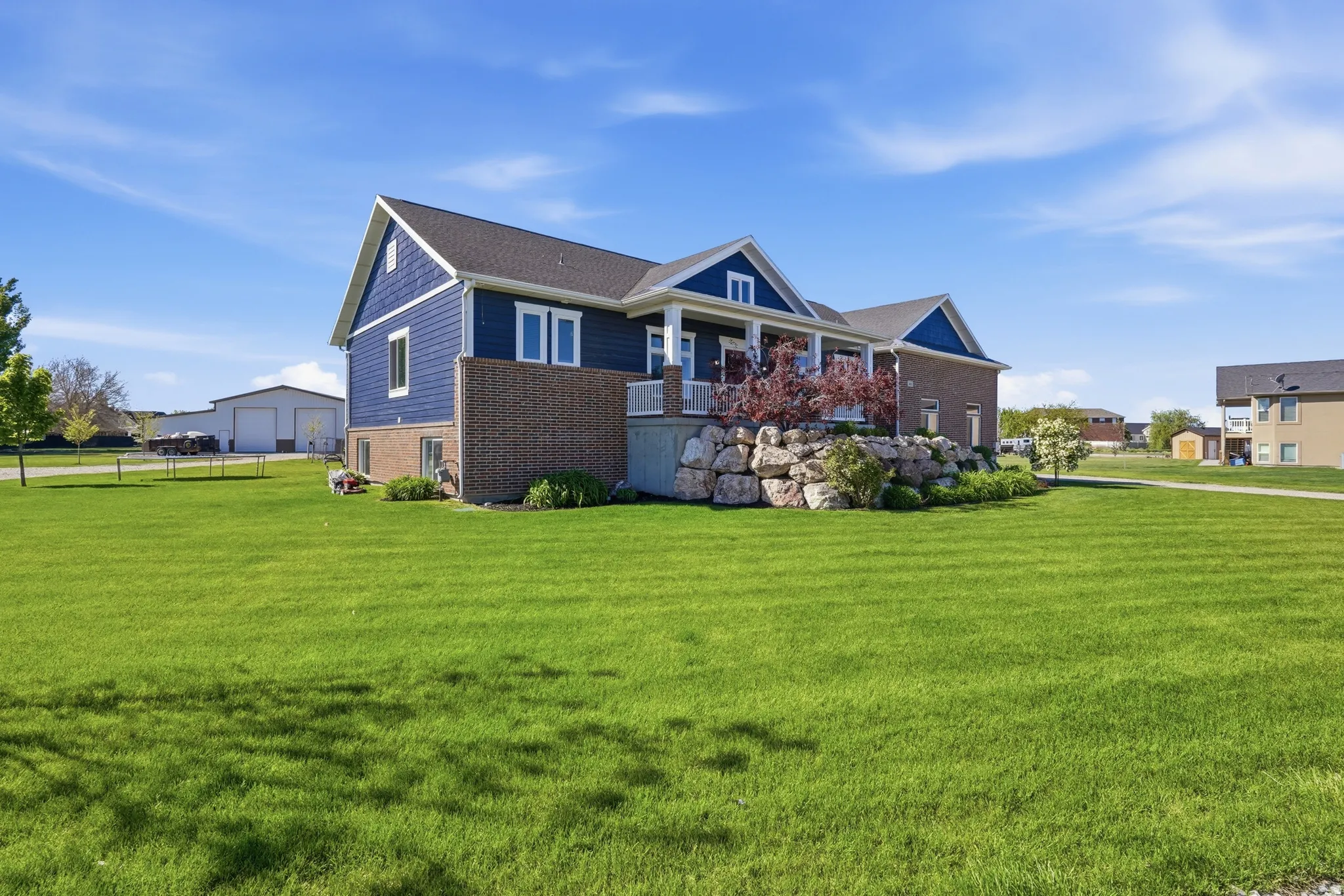 Spacious outdoor area featuring a manicured lawn, a blue siding exterior with brick accents, white trim, a covered porch, and a decorative rock retaining wall with established plantings