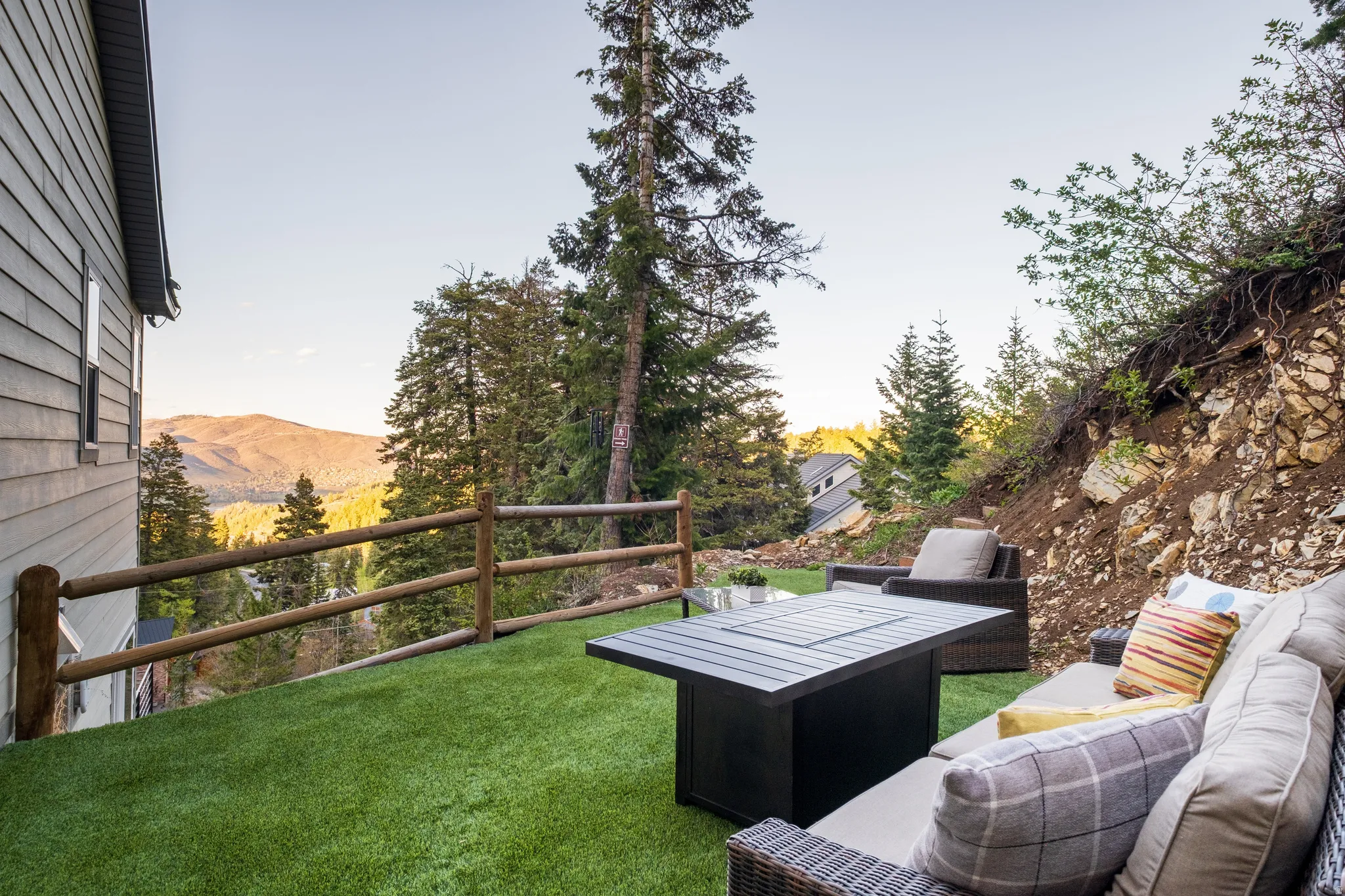 Outdoor living area featuring artificial turf, a rustic wooden rail fence, and a steep natural rock and earth hillside