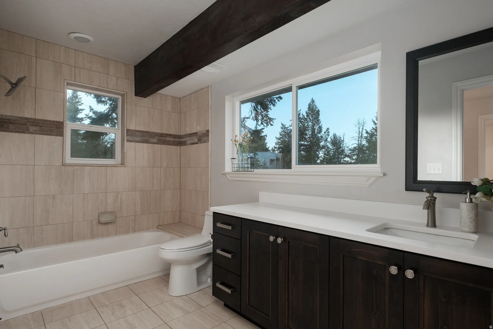 Bathroom featuring a white bathtub with tiled surround, dark wood-finish vanity with a white countertop, and a large window with natural light