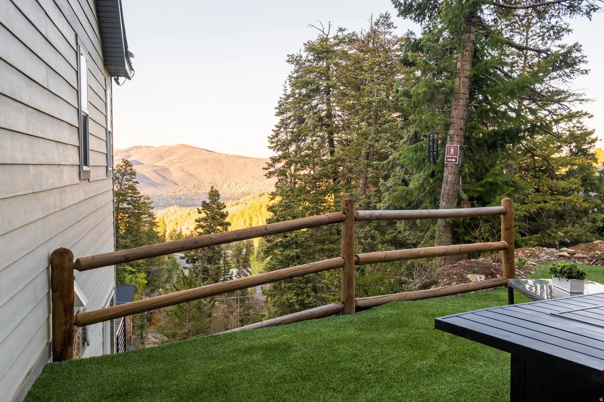 Siding exterior with multiple windows, featuring a natural wood split-rail fence, an artificial turf area, and a mountainous backdrop with abundant trees