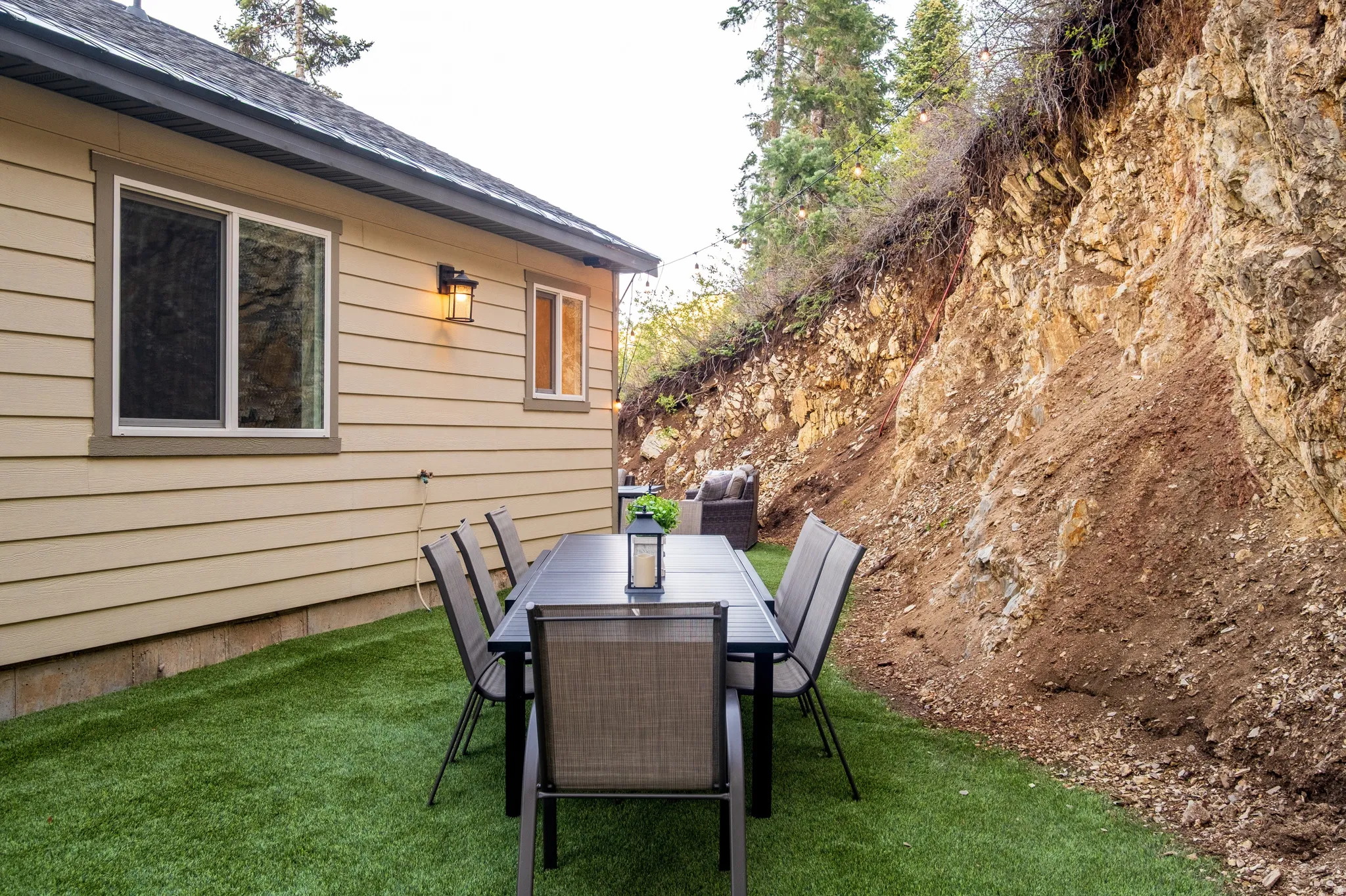 Outdoor dining area featuring artificial turf, a rock formation backdrop, exterior siding, and a wall-mounted light fixture