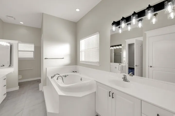 Spacious bathroom featuring a built-in jetted tub, dual vanities with white cabinetry, light countertops, and large format tile flooring