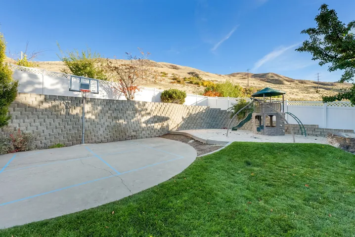 Expansive backyard featuring a concrete sport court with a basketball hoop, a sand play area, a multi-level playset with a slide, and a manicured lawn