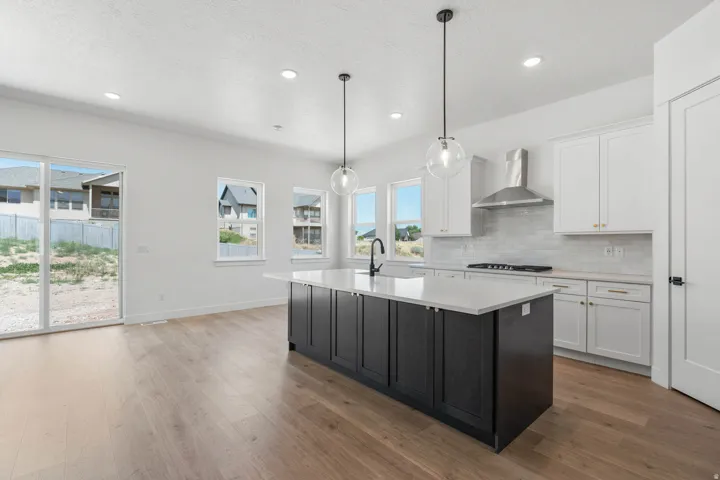 Kitchen featuring a dark-toned island with a white countertop, two pendant lights, wood-finish flooring, white cabinetry with gold-toned hardware, and a stainless steel range hood