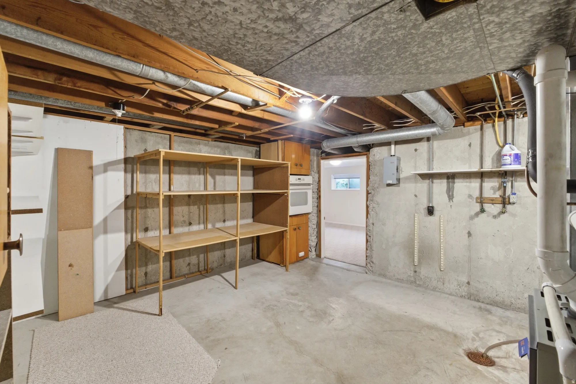 Unfinished basement featuring exposed wood ceiling joists, concrete flooring, built-in shelving, an installed wall oven, and a utility sink area