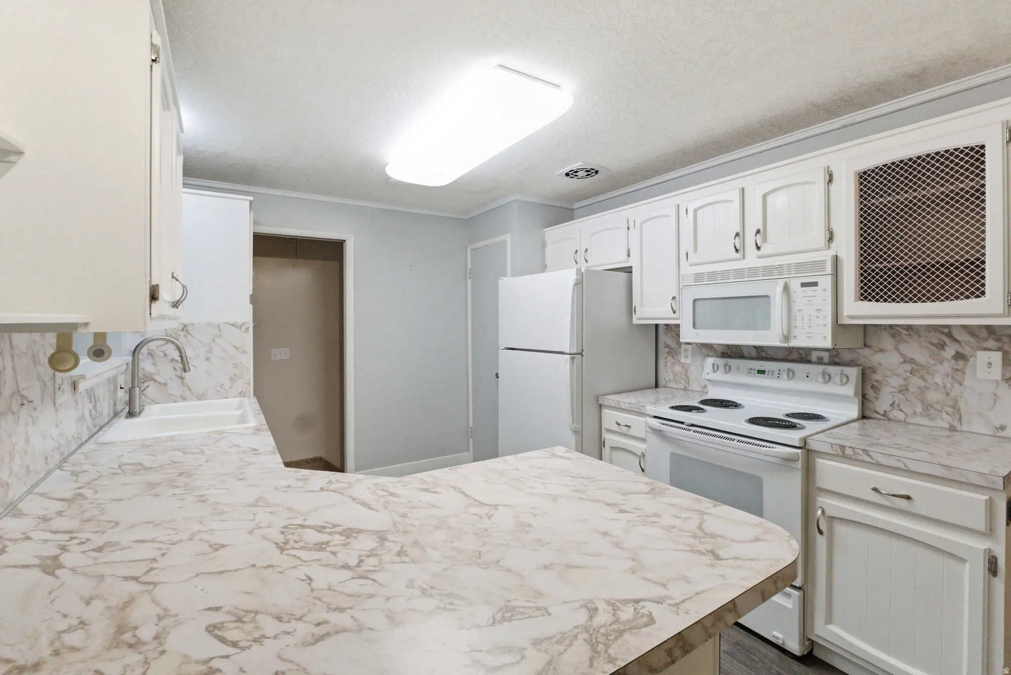 Kitchen featuring white cabinetry with brushed nickel hardware, white appliances, and light-toned, veined countertops extending to a breakfast bar