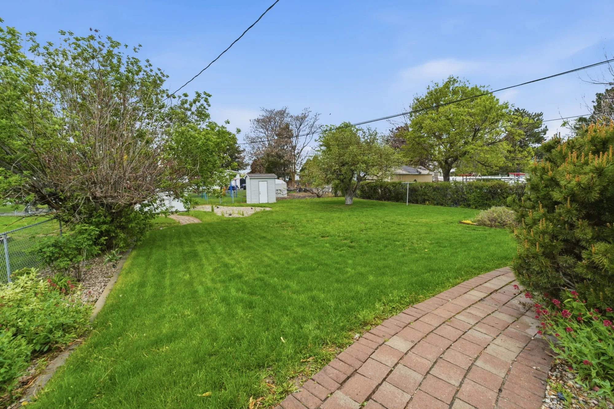 Expansive backyard featuring a manicured lawn, a brick paver pathway, mature trees, a chain-link fence, and a utility shed