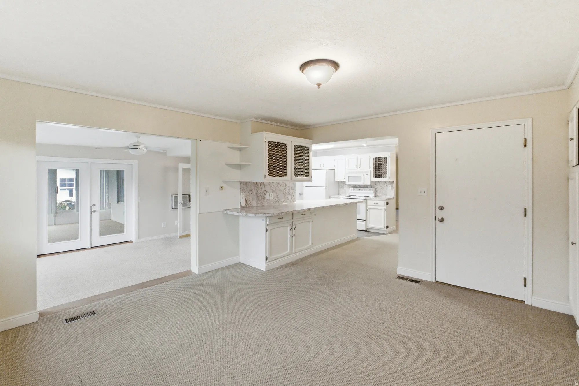 Open concept living area with light-colored carpeting, a kitchen bar with a patterned countertop, white cabinetry with glass-front uppers, and French patio doors opening to an additional room