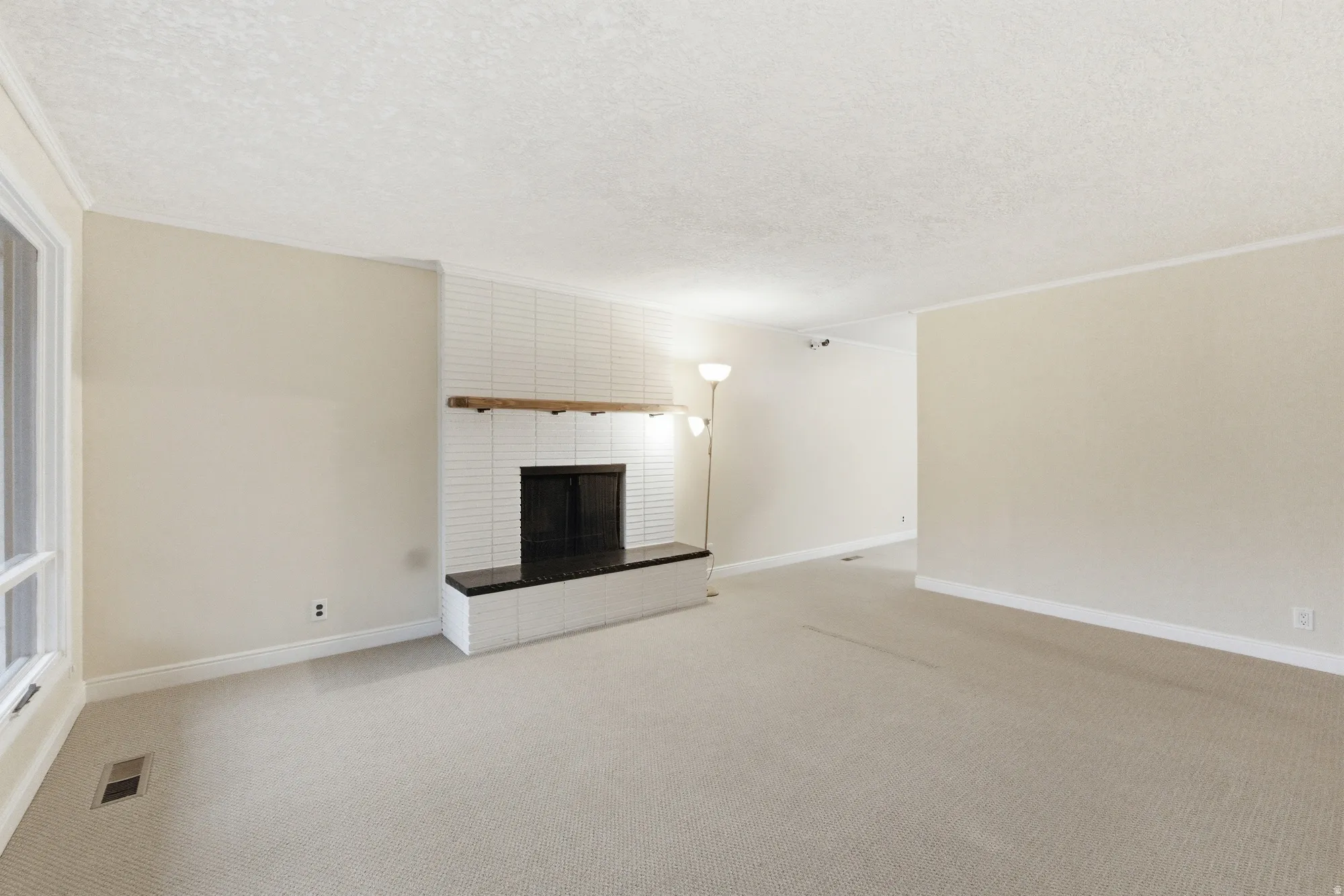 Living area featuring a white brick fireplace with a dark mantel, a wide window, light-toned carpeting, and crown molding