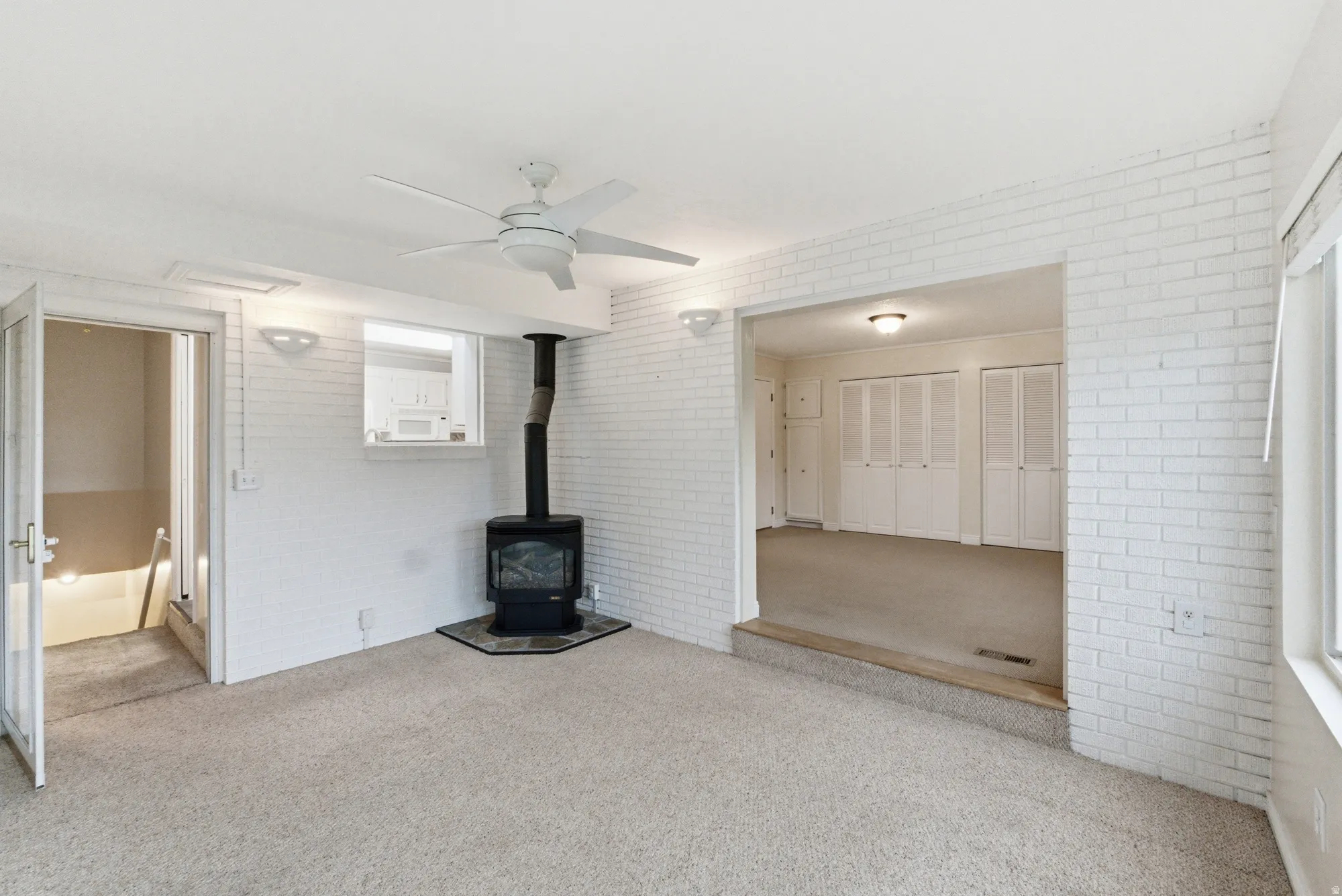 Carpeted room featuring a ceiling fan, a freestanding wood-burning stove with a stone hearth, white painted brick walls, and an open doorway to an additional room with bi-fold closet doors