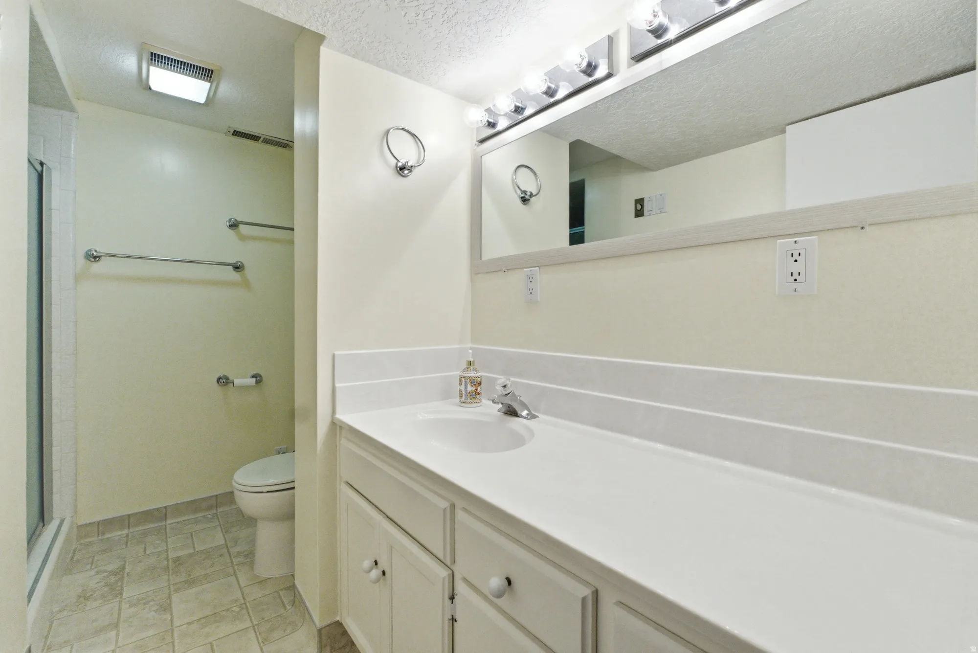 Bathroom featuring a white vanity with integrated sink, a large mirror with strip lighting, a commode, and a shower enclosure