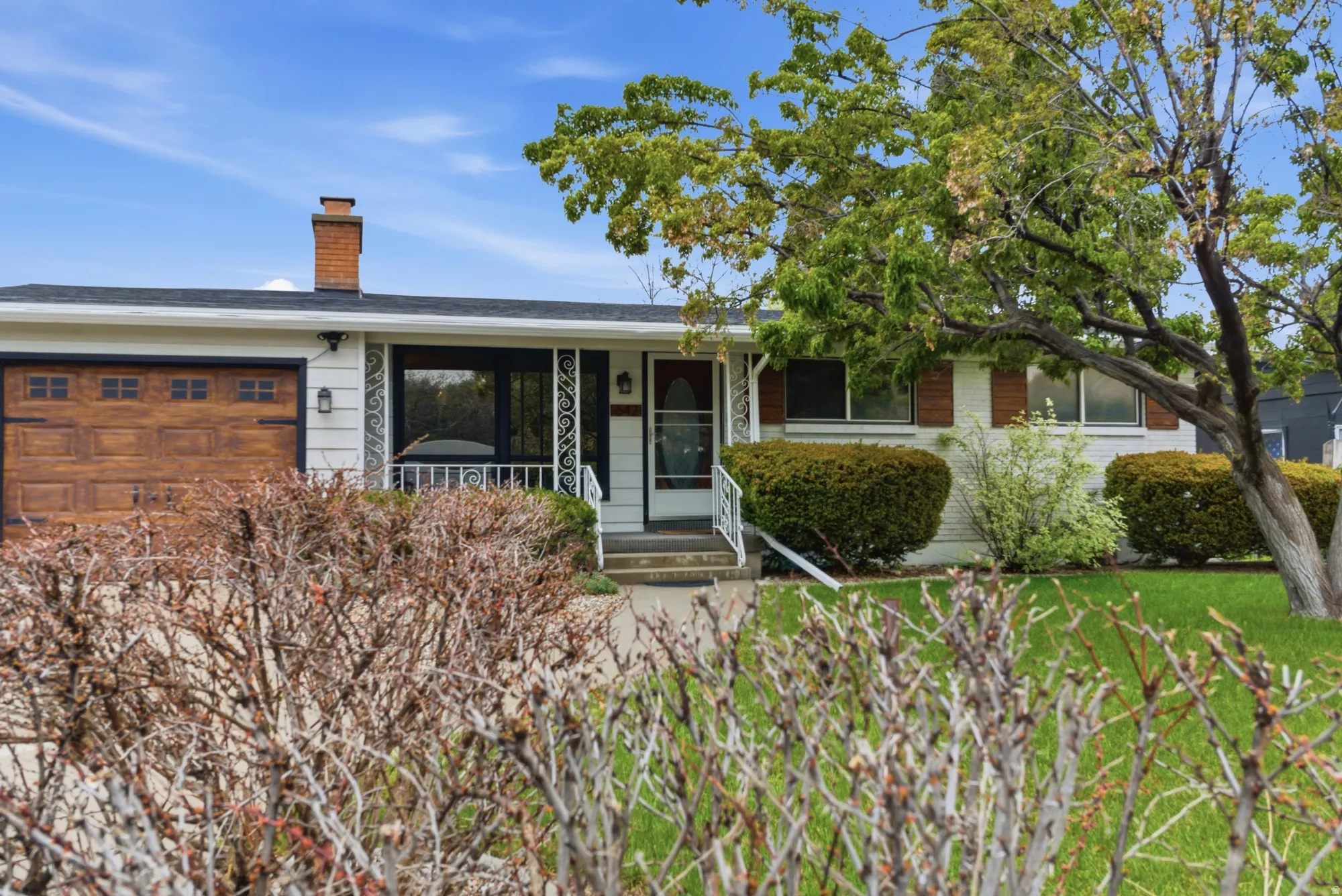 Single-story residence featuring a faux wood-finish garage door with window inserts, a front porch with decorative metal railings, and a prominent brick chimney