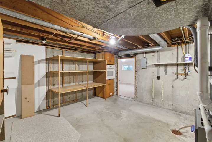 Unfinished basement featuring exposed wood ceiling joists, concrete flooring, built-in shelving, an installed wall oven, and a utility sink area