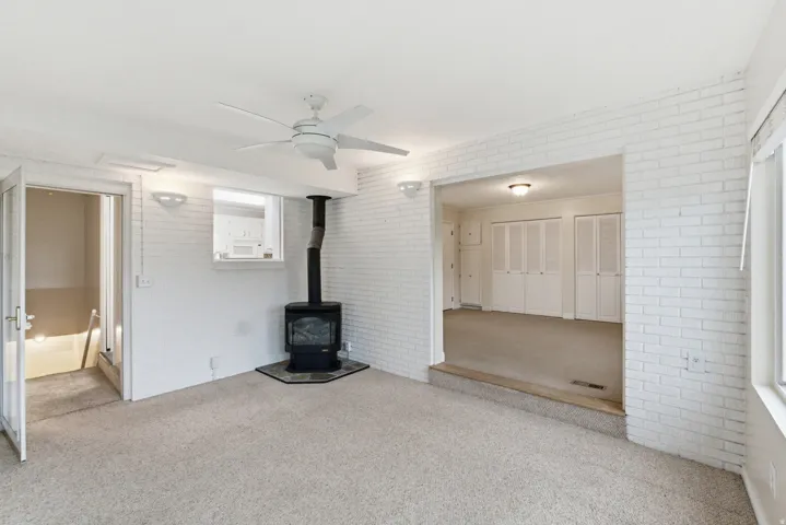 Carpeted room featuring a ceiling fan, a freestanding wood-burning stove with a stone hearth, white painted brick walls, and an open doorway to an additional room with bi-fold closet doors