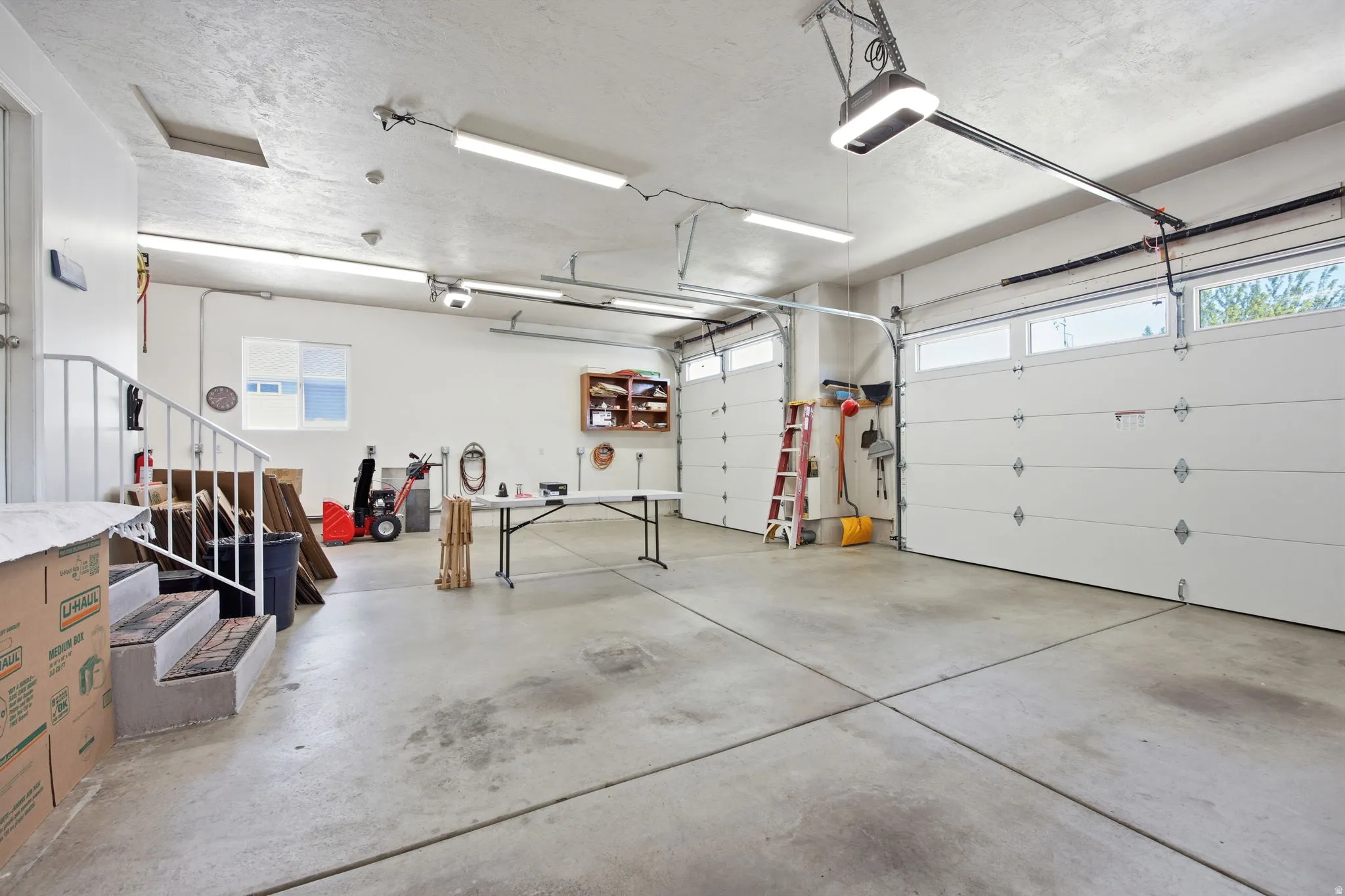 Spacious two-car garage featuring a concrete floor, multiple fluorescent light fixtures, two overhead garage doors with window panels, and a built-in shelving unit