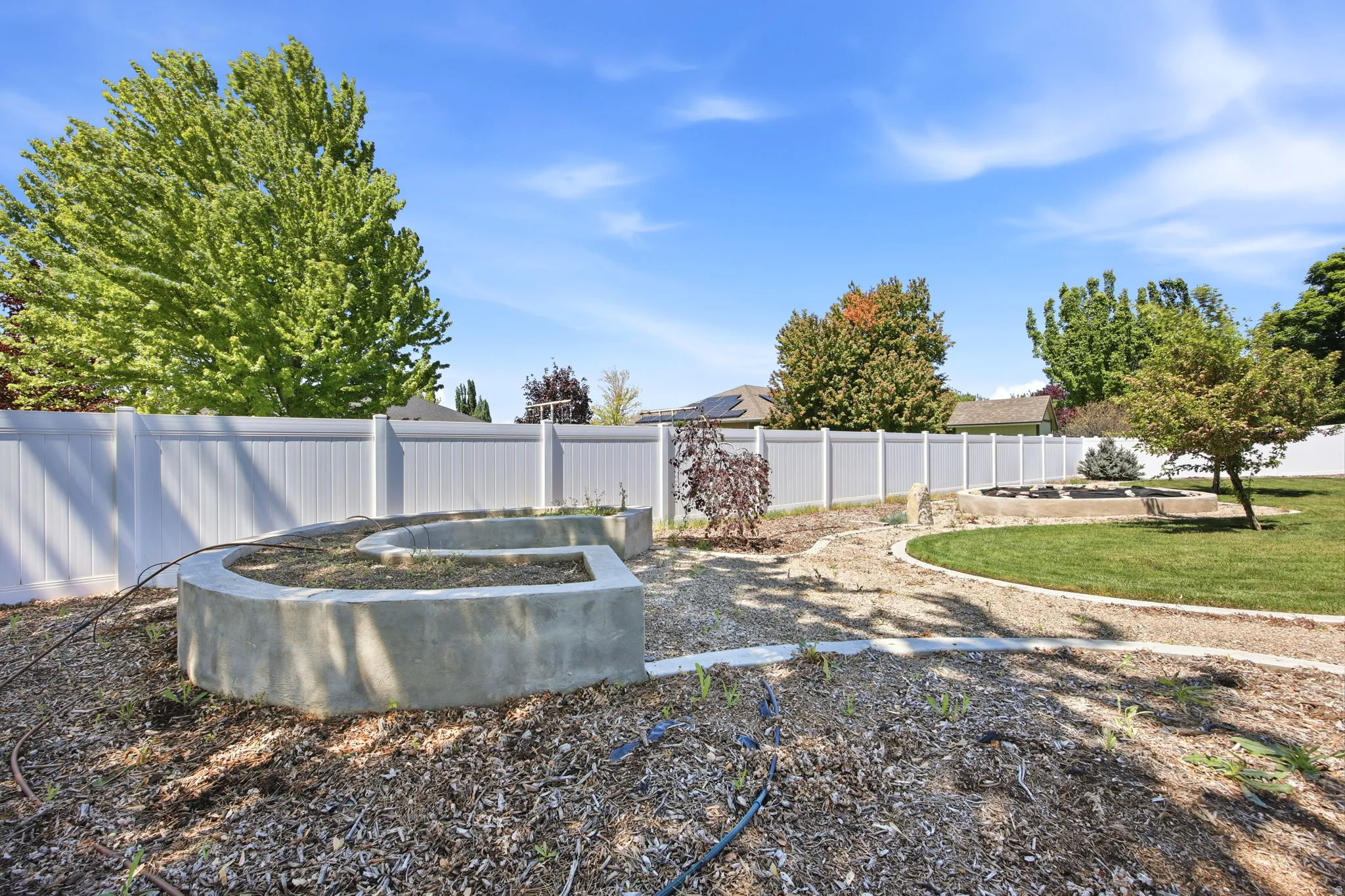 Expansive backyard featuring a white vinyl privacy fence, concrete raised garden beds, a gravel path, mature trees, and a lawn area