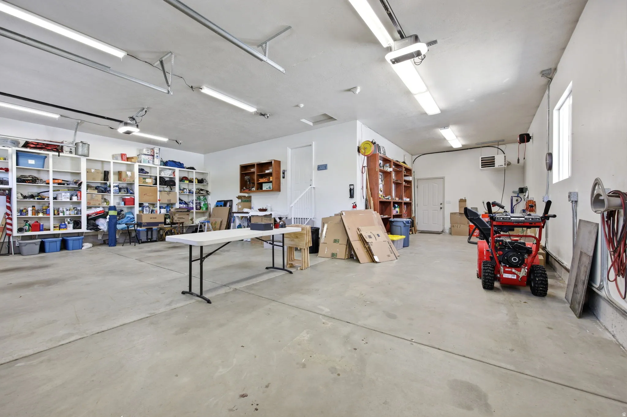 Spacious garage featuring a concrete floor, built-in shelving, ample overhead lighting, multiple windows, and a dedicated utility door