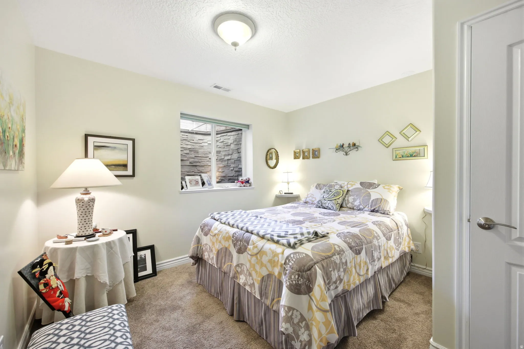 Bedroom featuring a window with a stone-look exterior, neutral-toned walls, and textured carpet flooring