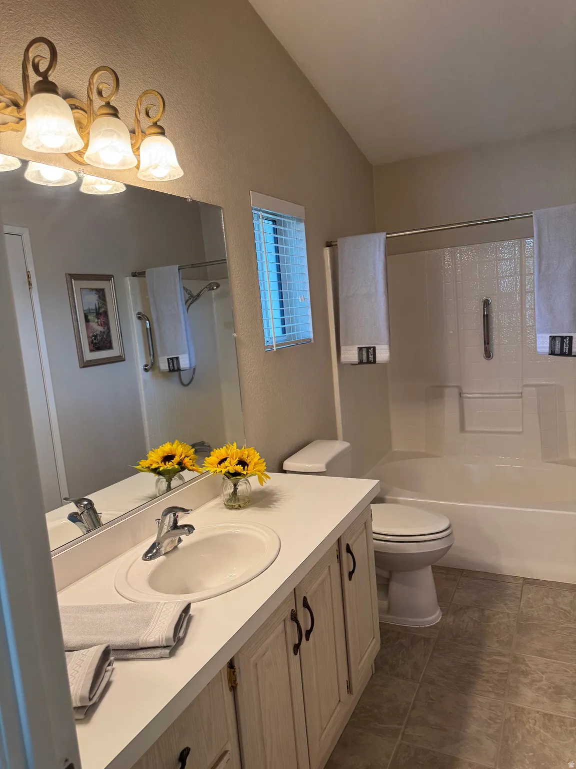 Bathroom featuring a single vanity with an integrated sink, an overhead vanity light fixture, a mirrored wall, a window with horizontal blinds, a toilet, and a combination tub and shower with a textured surround