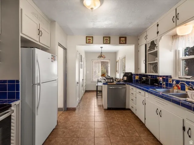 Galley kitchen featuring white cabinetry with decorative upper accents, blue tiled countertops and backsplash, tile flooring, and a stainless steel dishwasher