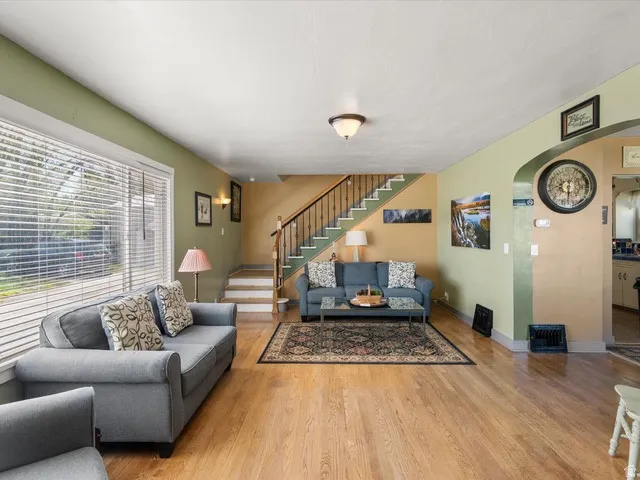 Spacious living area featuring wood-finish flooring, a large picture window with blinds, and a staircase with wrought iron balusters