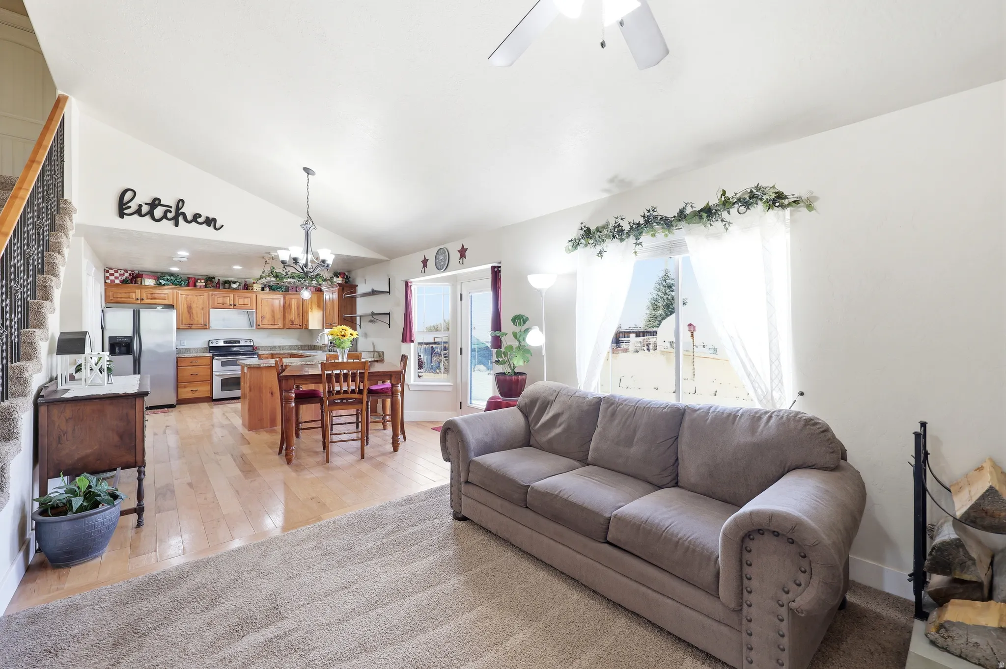 Open-concept living area featuring vaulted ceilings, light wood-finish flooring, a kitchen with wood cabinetry and stainless steel appliances, and a carpeted living space with a large window