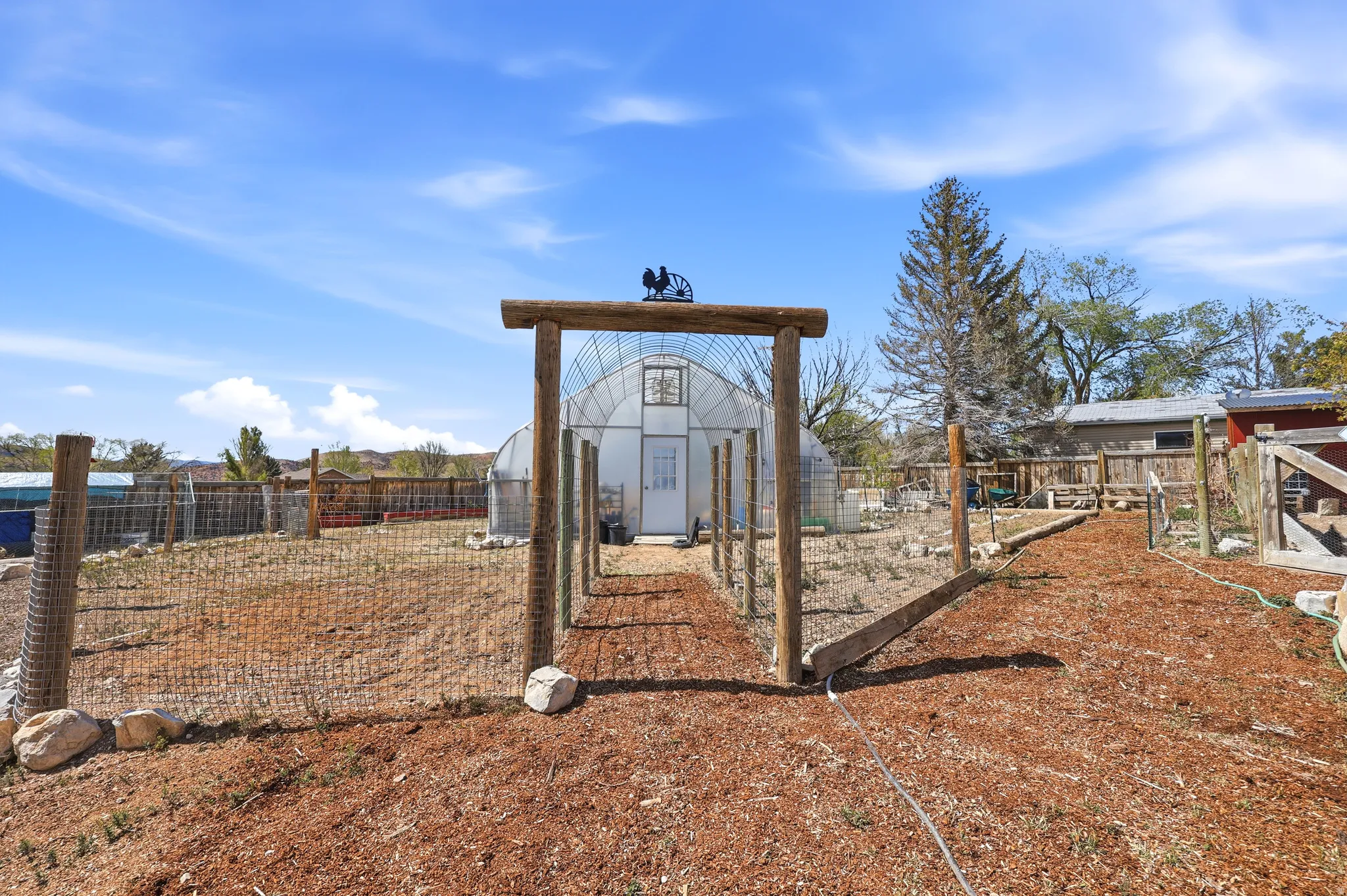 Spacious outdoor area featuring a prominent greenhouse structure, a rustic wooden entry arch, and perimeter fencing
