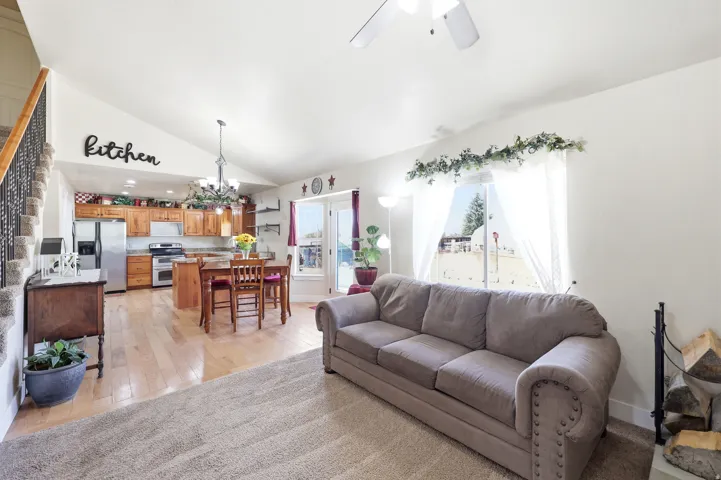Open-concept living area featuring vaulted ceilings, light wood-finish flooring, a kitchen with wood cabinetry and stainless steel appliances, and a carpeted living space with a large window
