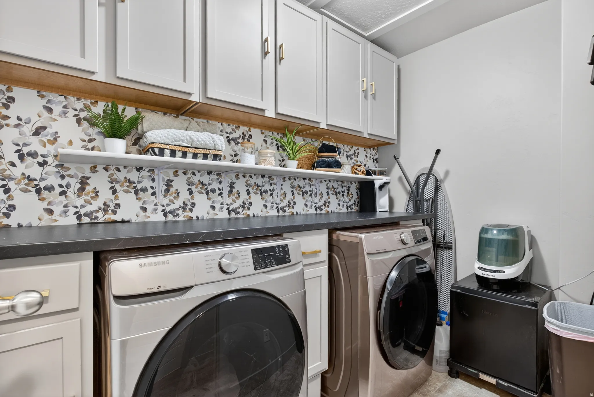 Laundry area featuring a dark countertop, white upper cabinetry with gold hardware, and a decorative floral backsplash