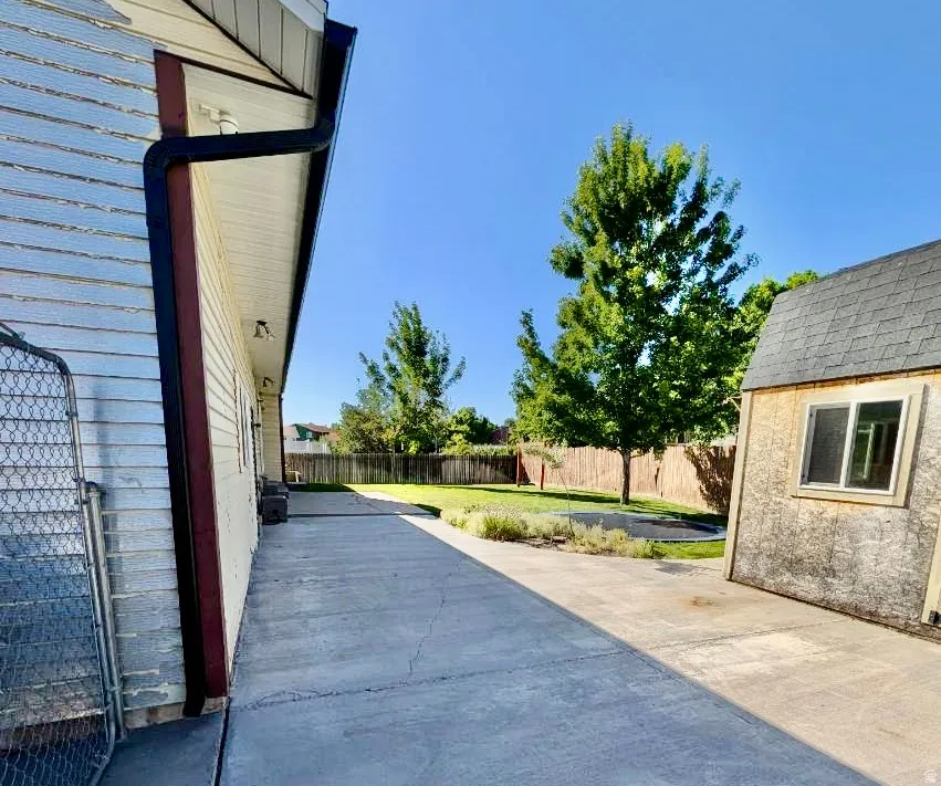 Expansive paved area featuring a detached storage shed with shingled roof, a wooden privacy fence, mature trees, and a grassy yard