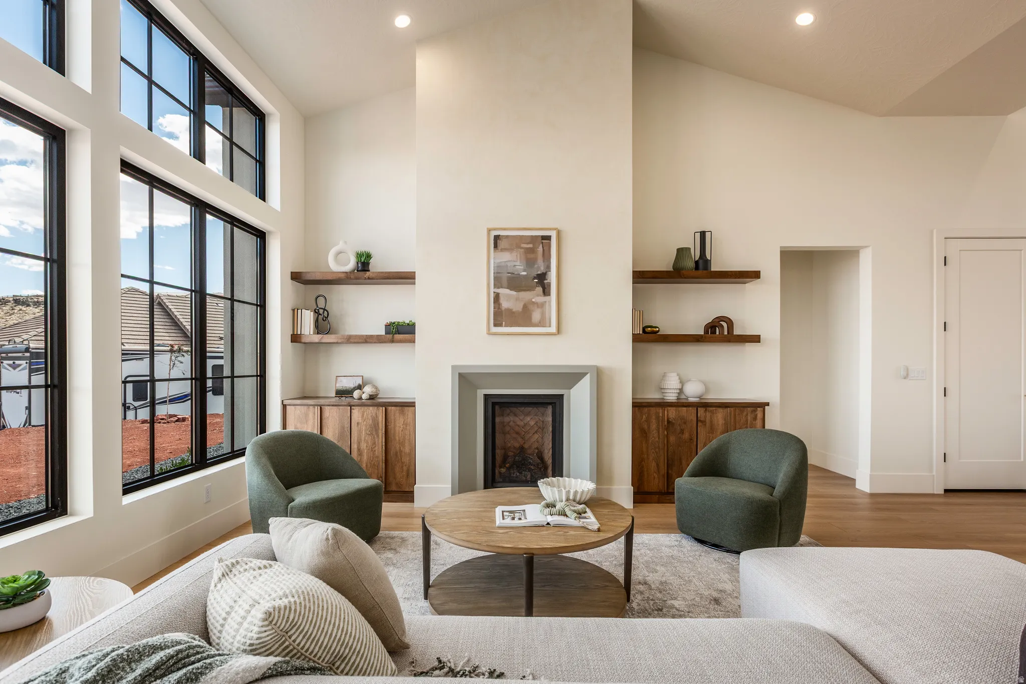 Vaulted ceiling living area featuring a central fireplace with a light-toned stone surround, integrated wood cabinetry, floating wood shelves, and large grid-pattern windows