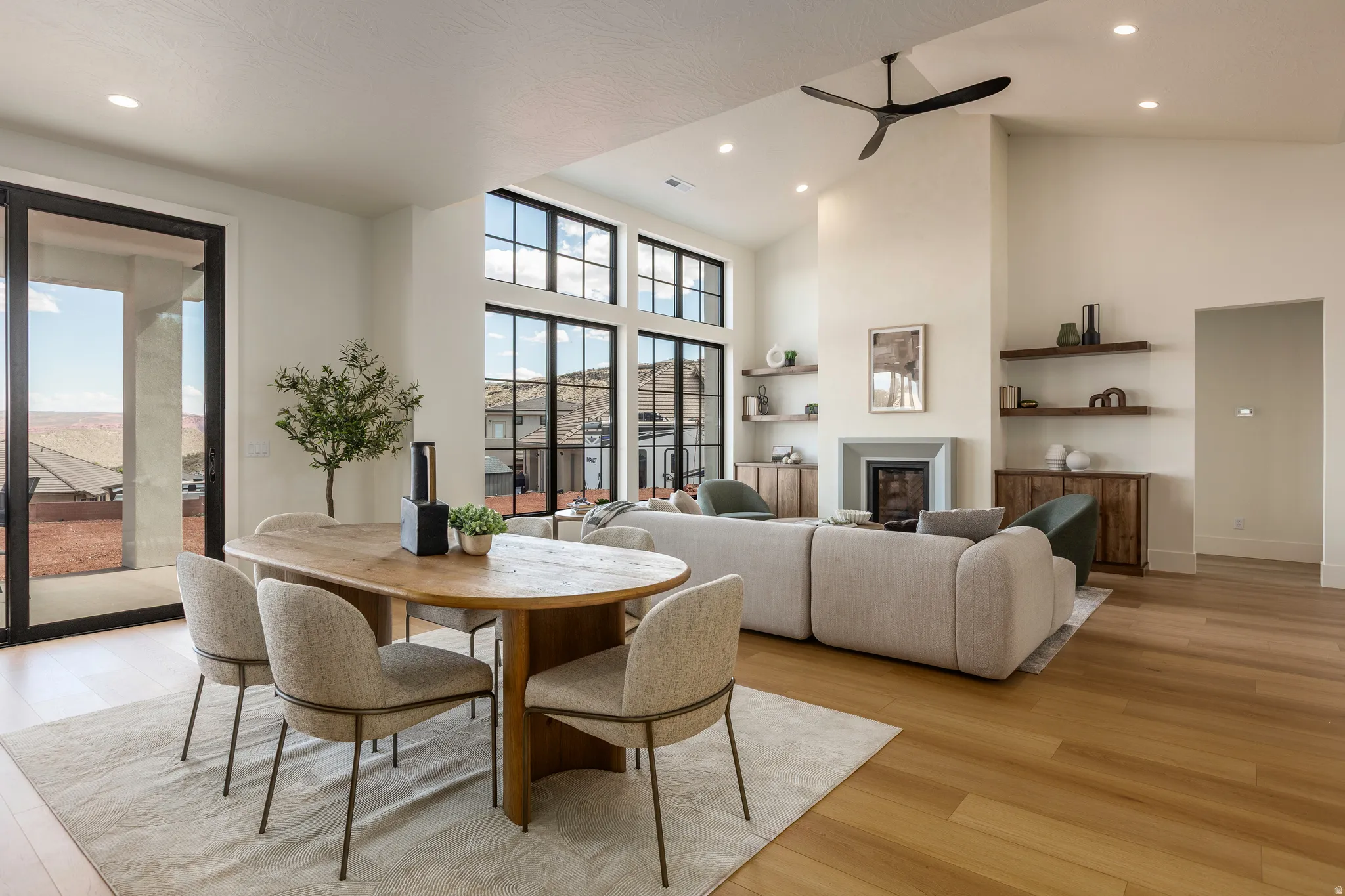 Open-concept living area featuring high vaulted ceilings, expansive black-framed windows, wood-finish flooring, a contemporary fireplace with a stone surround, and recessed lighting