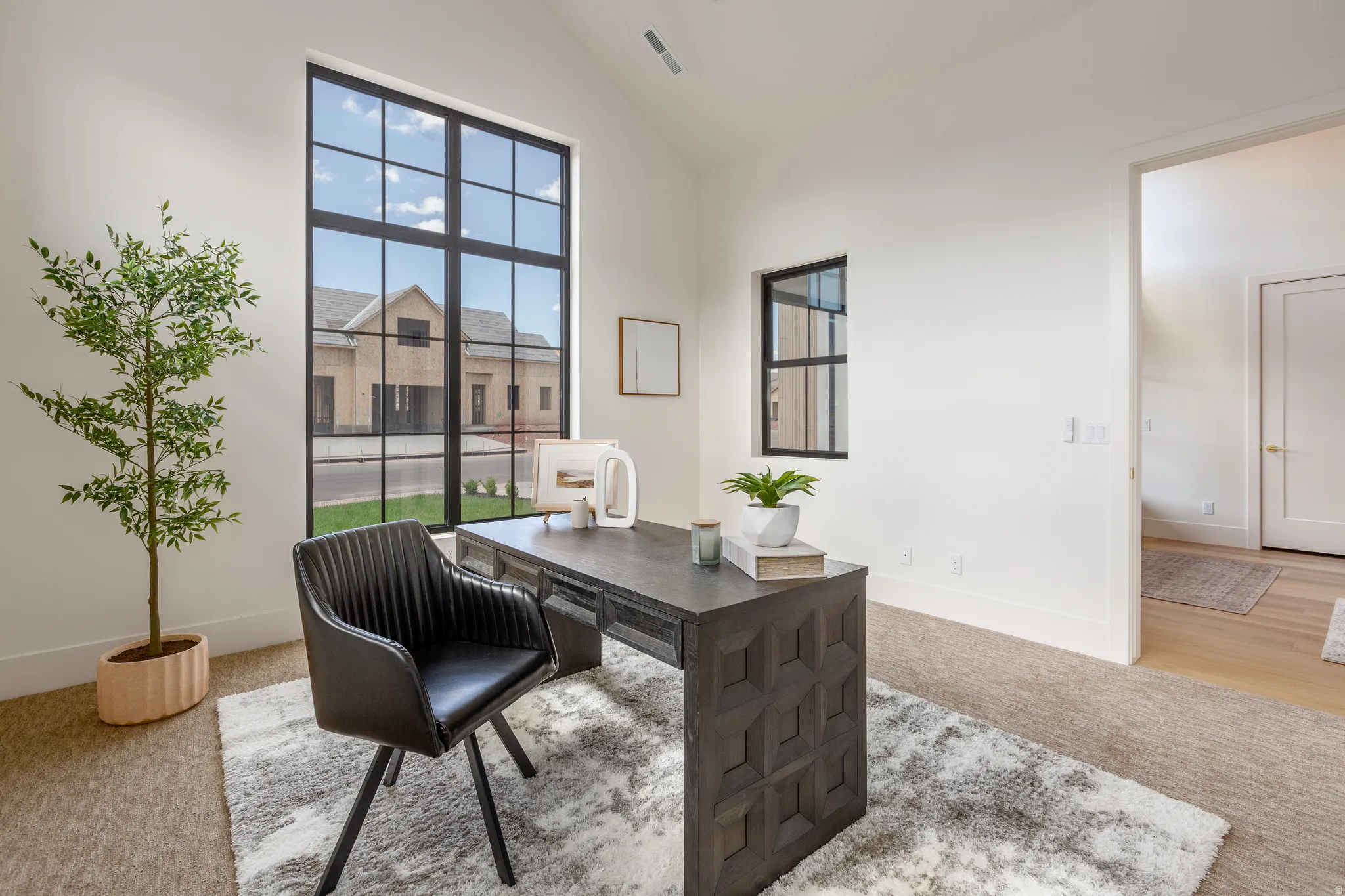 Spacious interior room featuring high ceilings, large grid-pattern windows, light-tone carpeting, and an open doorway to a hard-surface flooring area